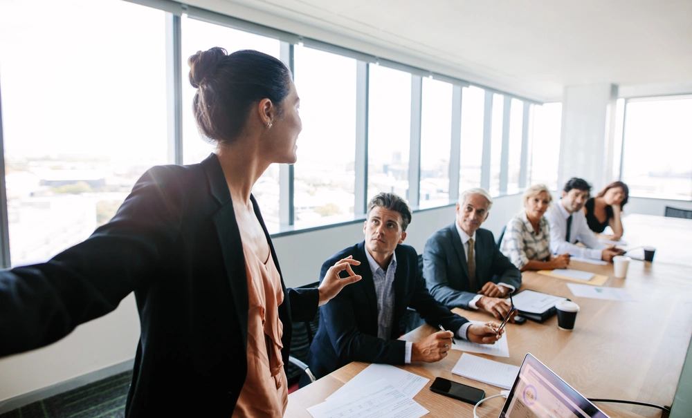 © Jacob Lund / Fotolia Eine Frau in Bürokleidung spricht zu 5 Zuhörern an einem Meetingtisch.